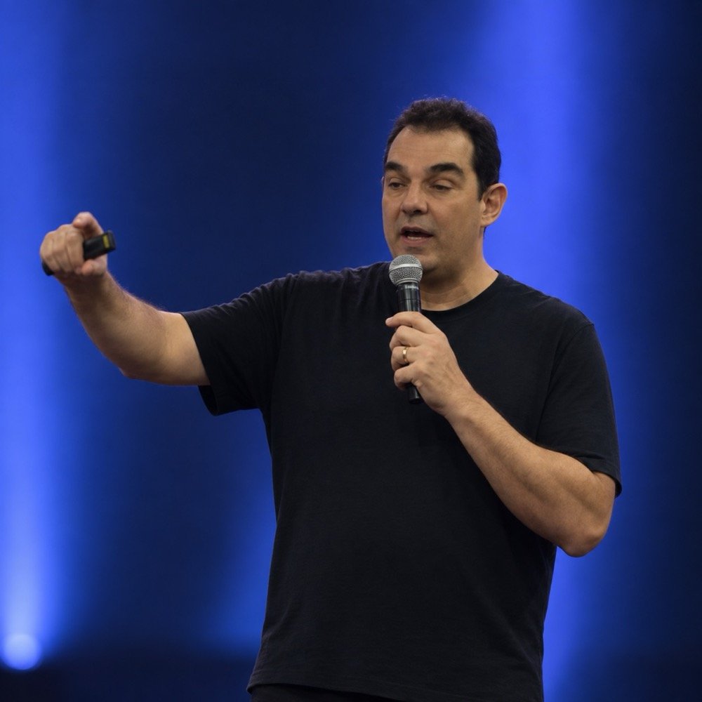 Victor Mirshawka Jr. palestrando, com microfone e controle de apresentação, sob luz azul de palco